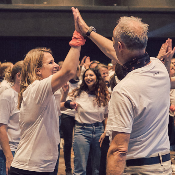 Two people smiling and giving a high five in a group setting with others clapping in the background.