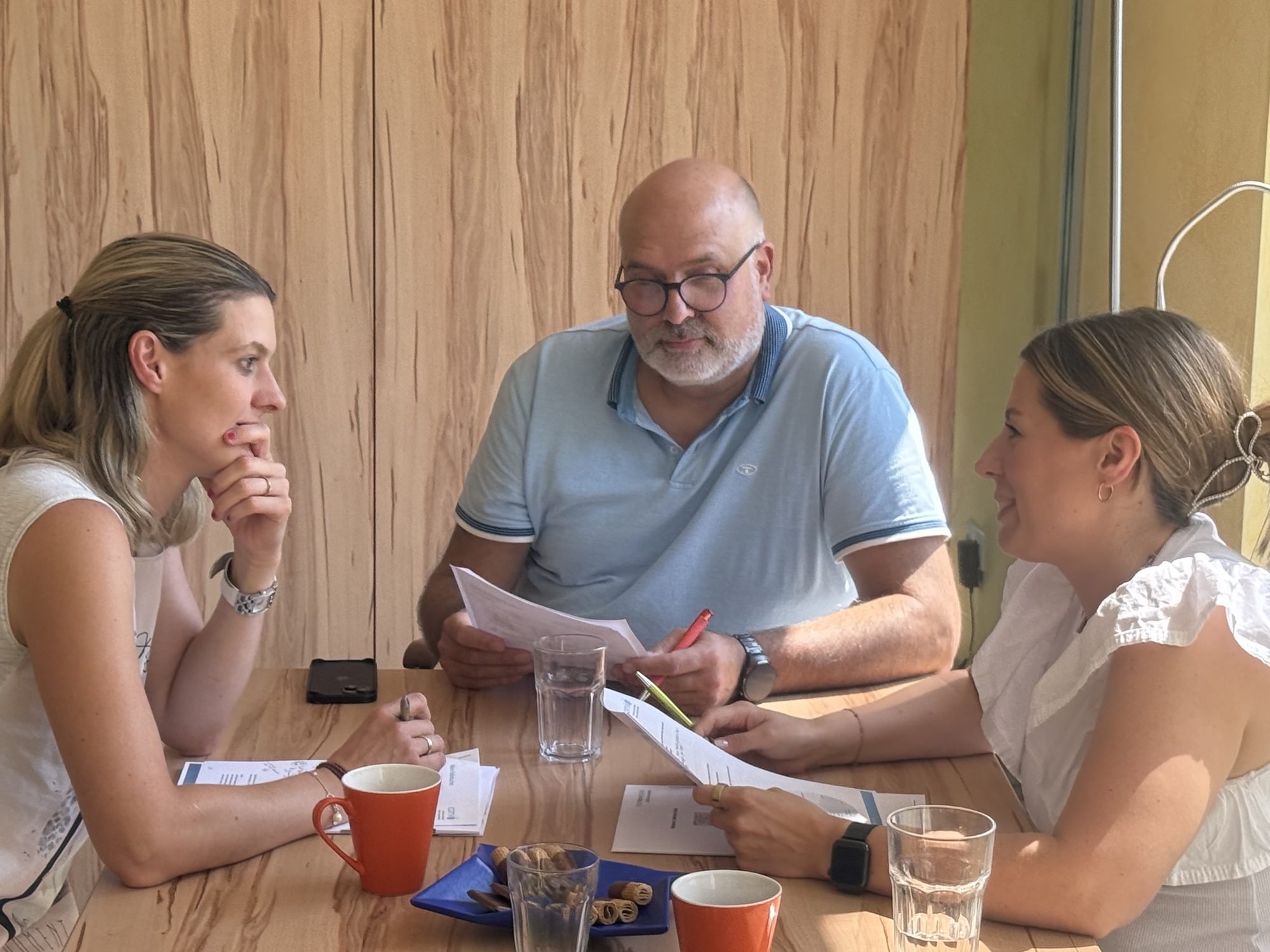 Drei Teilnehmende arbeiten am Tisch vor der Holzwand