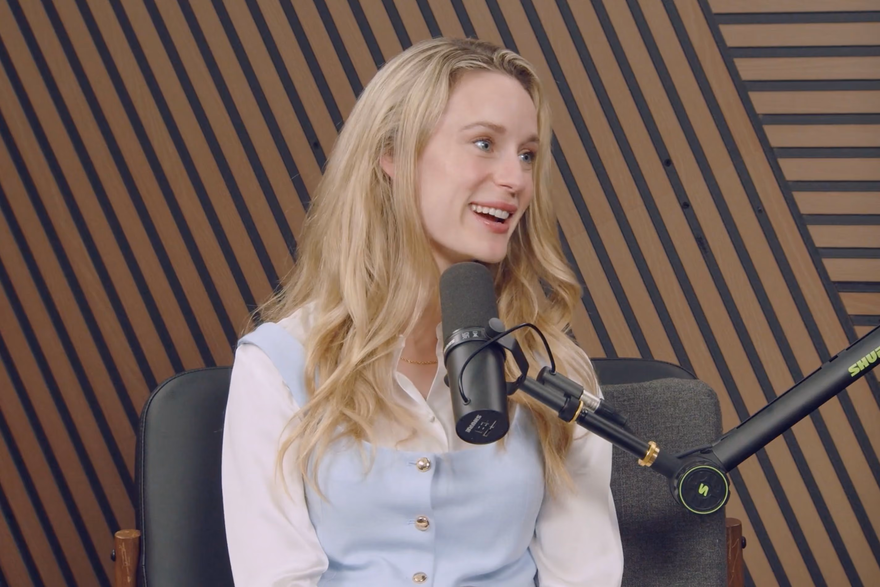 Smiling blonde woman speaking into a microphone in a modern podcast studio with wood paneling.