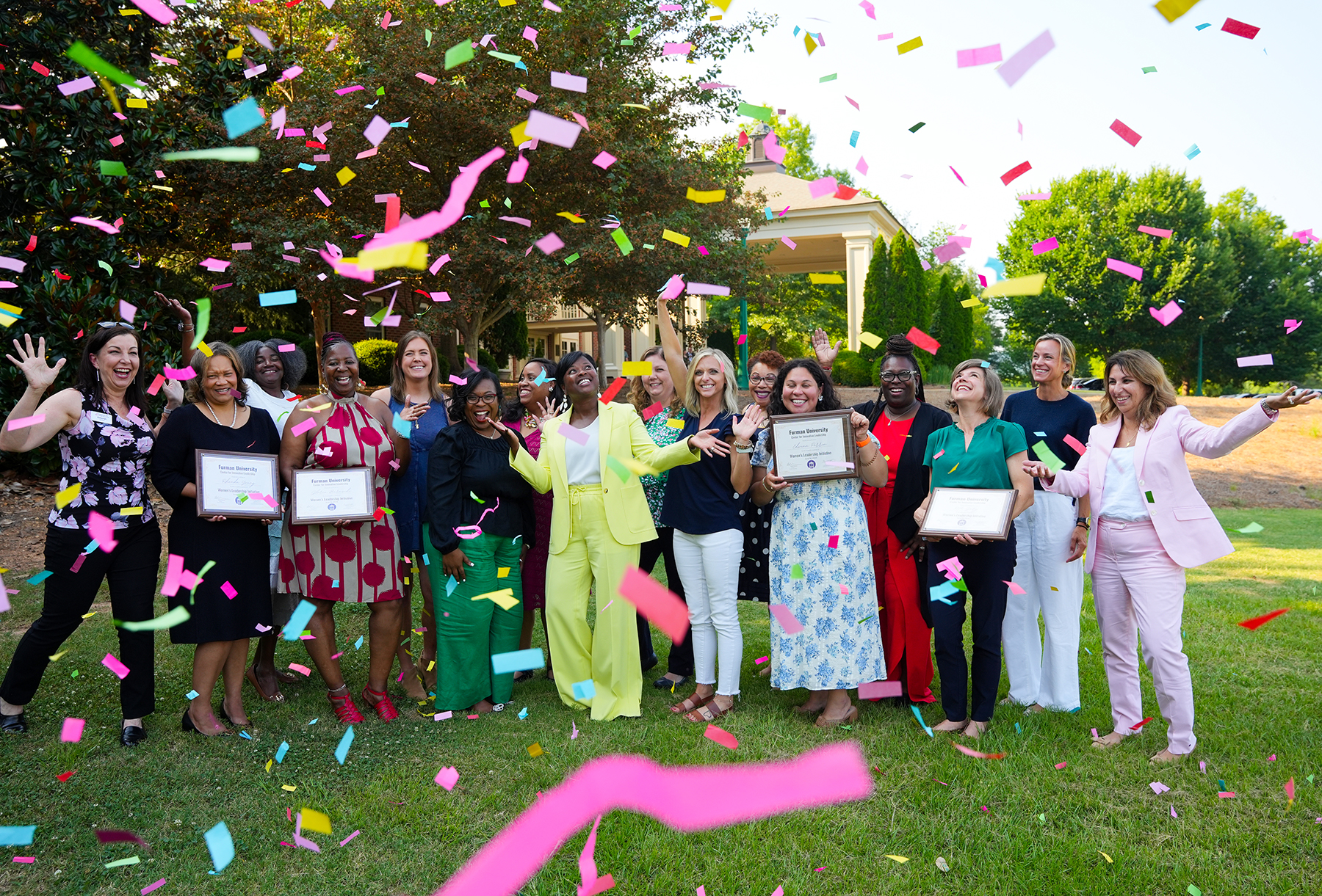 A joyful group of women stands outdoors at Furman University, celebrating their Women's Leadership Initiative graduation with certificates in hand as colorful confetti fills the air.