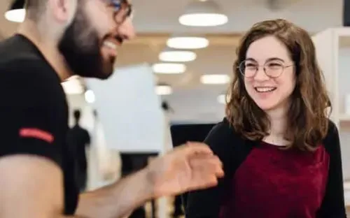 A male student and a female student share a laugh during a conversation in a bright workspace.