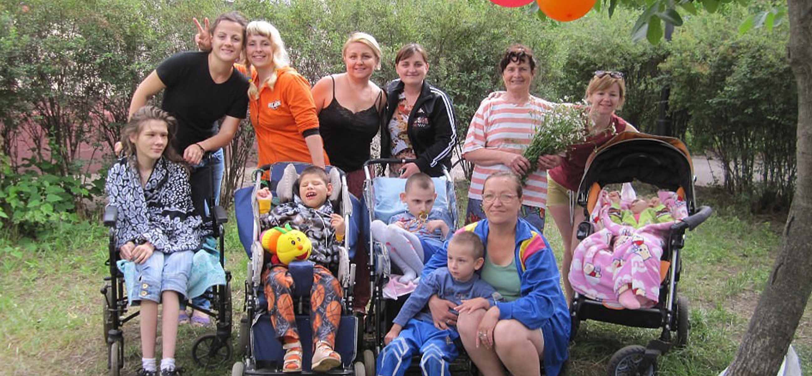 Mothers and caregivers gather outdoors with their children, several of whom sit in wheelchairs and adaptive strollers, beneath red and orange balloons.