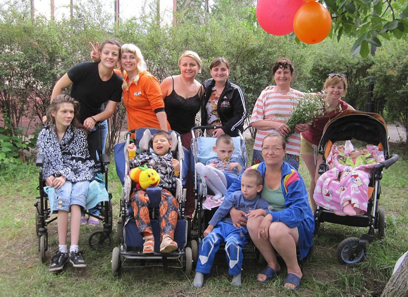 Mothers and caregivers gather outdoors with their children, several of whom sit in wheelchairs and adaptive strollers, beneath red and orange balloons.