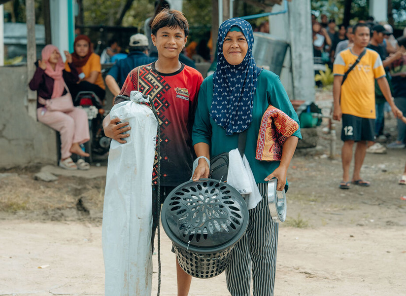 A smiling teenage boy and a woman in a headscarf carry shelter supplies, including a tarp and a cooking pot, through a busy crowd.