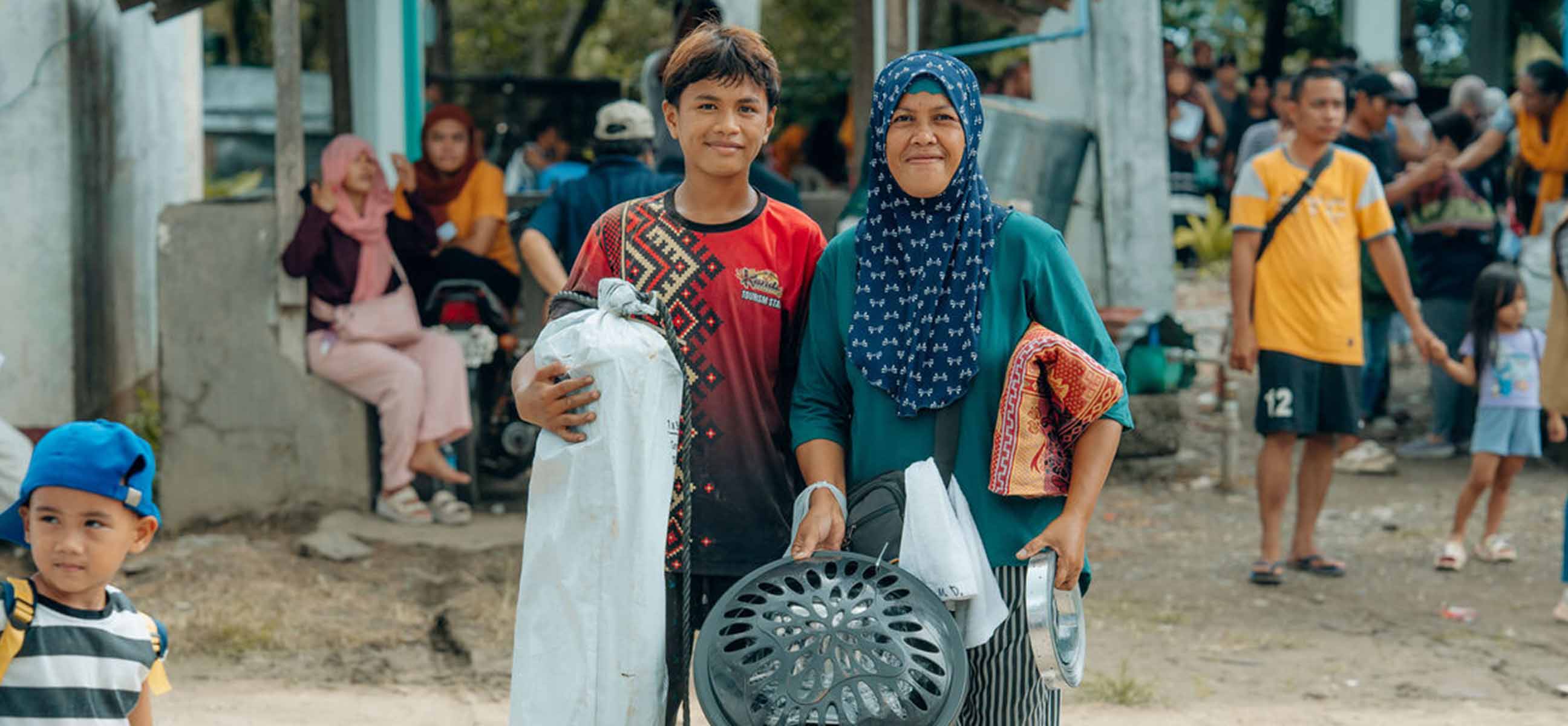 A smiling teenage boy and a woman in a headscarf carry shelter supplies, including a tarp and a cooking pot, through a busy crowd.