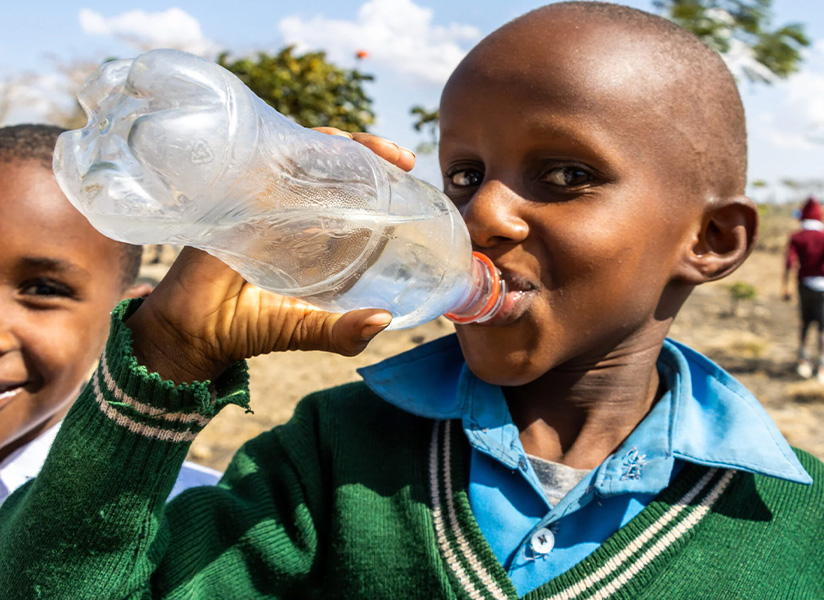 A schoolboy in a green sweater drinks clean water from a plastic bottle while a smiling classmate looks on.