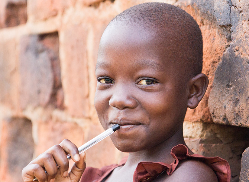 A young girl leans against a brick wall, smiling shyly with a pen resting at her lips.