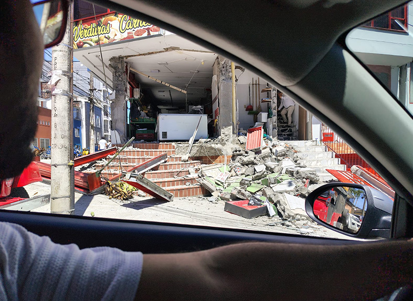 Seen through a car window, a storefront lies collapsed into a pile of rubble on a city street.