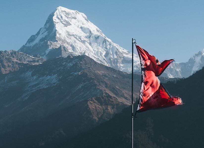 The flag of Nepal flies in front of a snow-capped Himalayan peak.