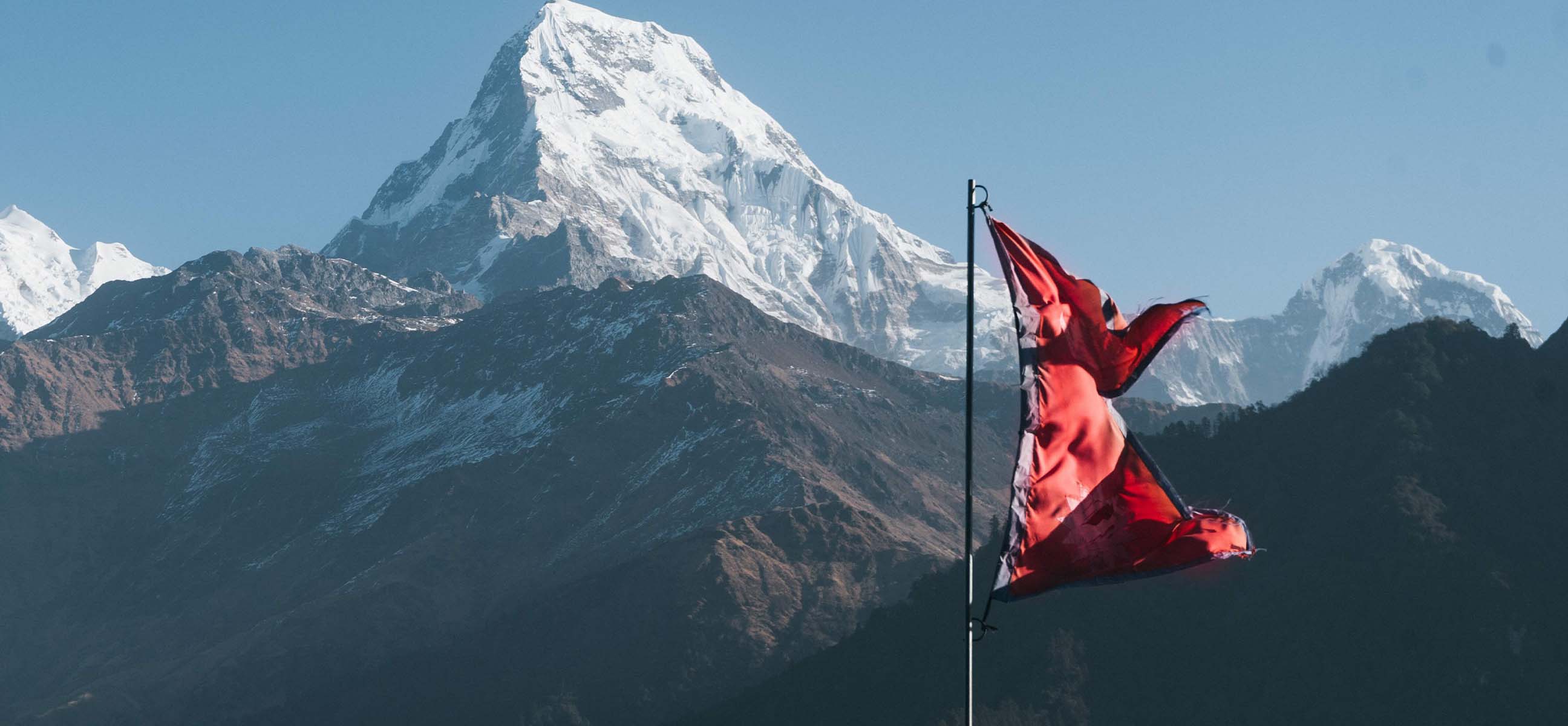 The flag of Nepal flies in front of a snow-capped Himalayan peak.