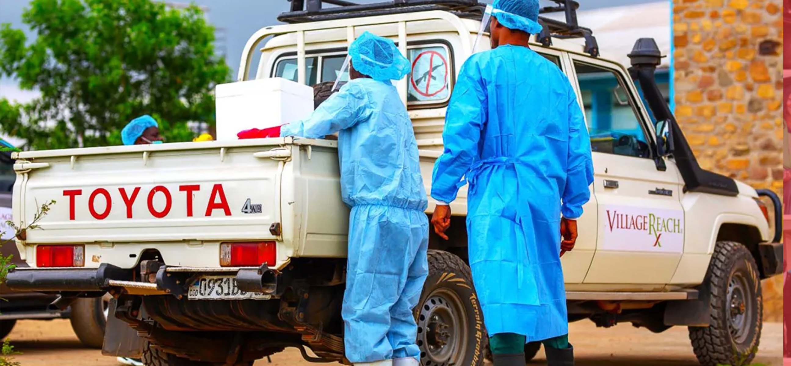 Health workers in blue protective gowns and caps unload supplies from the back of a white pickup truck.