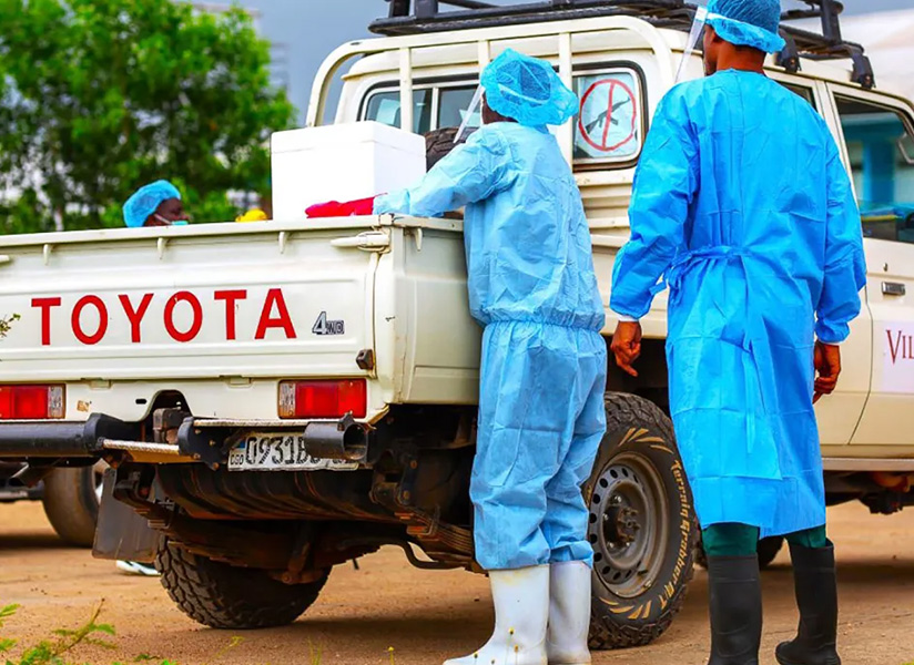 Health workers in blue protective gowns and caps unload supplies from the back of a white pickup truck.