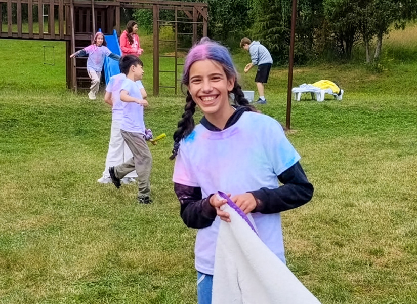 A smiling girl with a purple headband, holding a white sheet, plays a game on a grassy lawn with other children at summer camp.