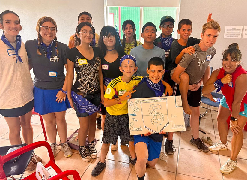 Smiling teenagers and volunteers in blue bandanas crowd together around a hand-drawn poster.