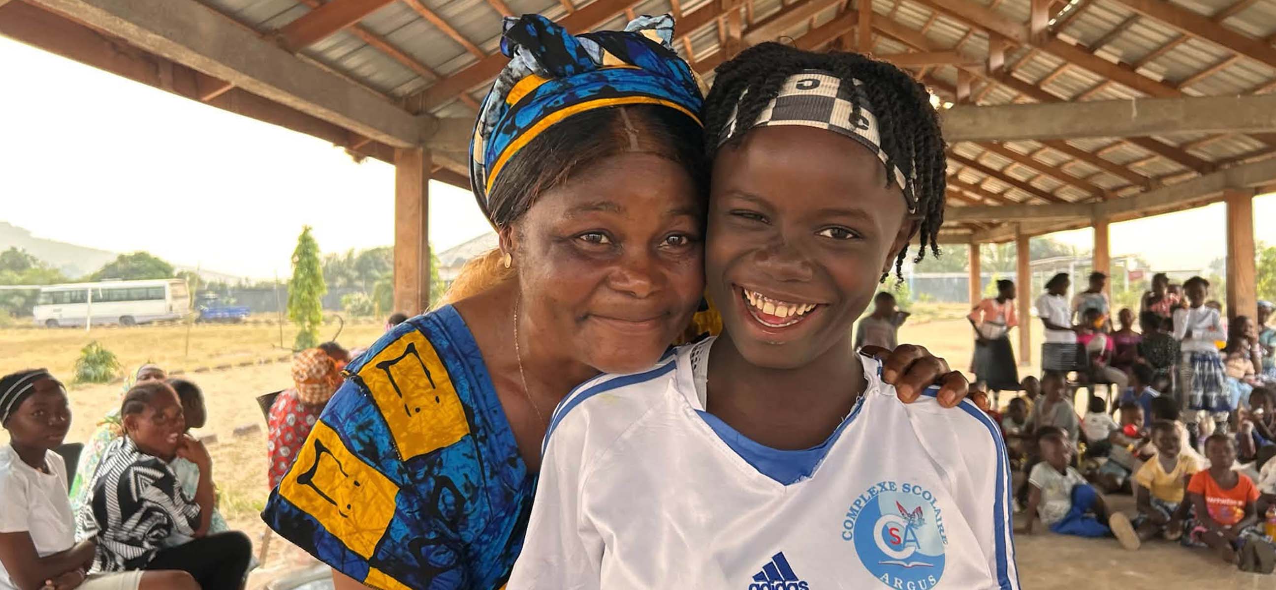 A foster mother in a colorful headwrap hugs a beaming girl under an open-air pavilion filled with children.