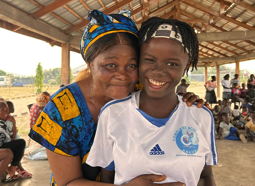 A foster mother in a colorful headwrap hugs a beaming girl under an open-air pavilion filled with children.