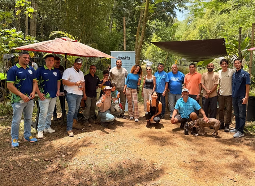 A large group of farmers and Asekia staff pose together outdoors beneath trees and a patio umbrella.