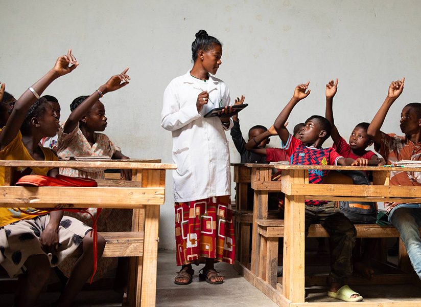 A teacher in a white coat stands among wooden desks as her students eagerly raise their hands.