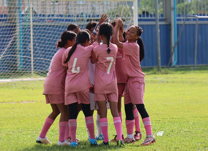 Girls in pink football uniforms huddle together to celebrate on a grass pitch.