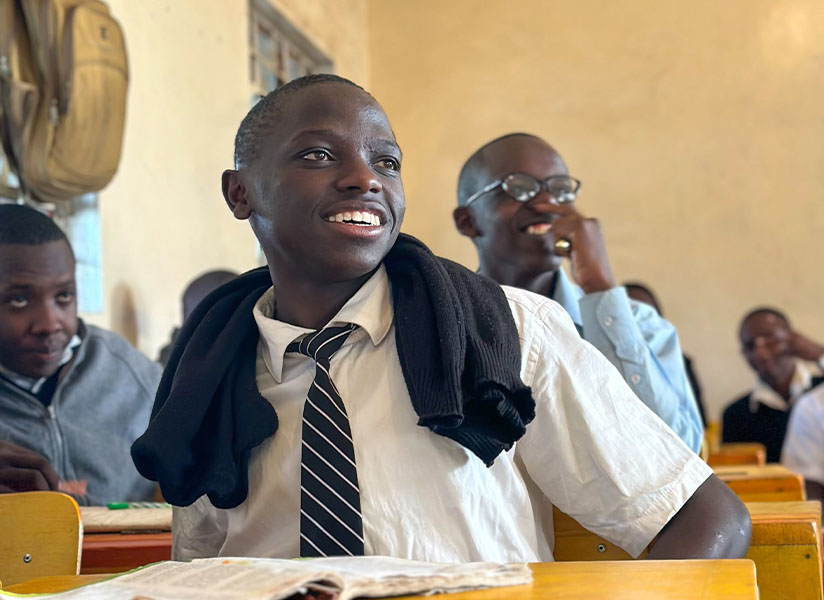 A smiling student in a white shirt and striped tie sits in class, a sweater draped over his shoulders.