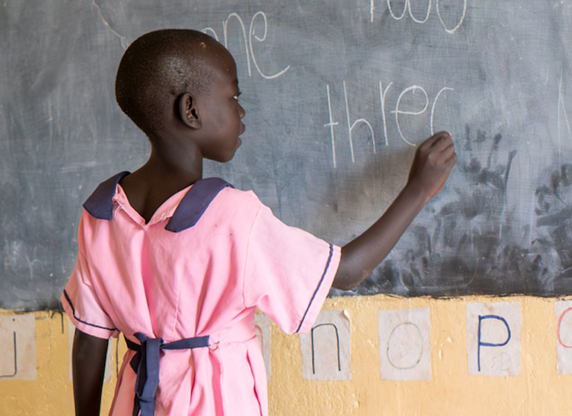 A young girl in a pink school uniform writes words in chalk on a classroom blackboard.