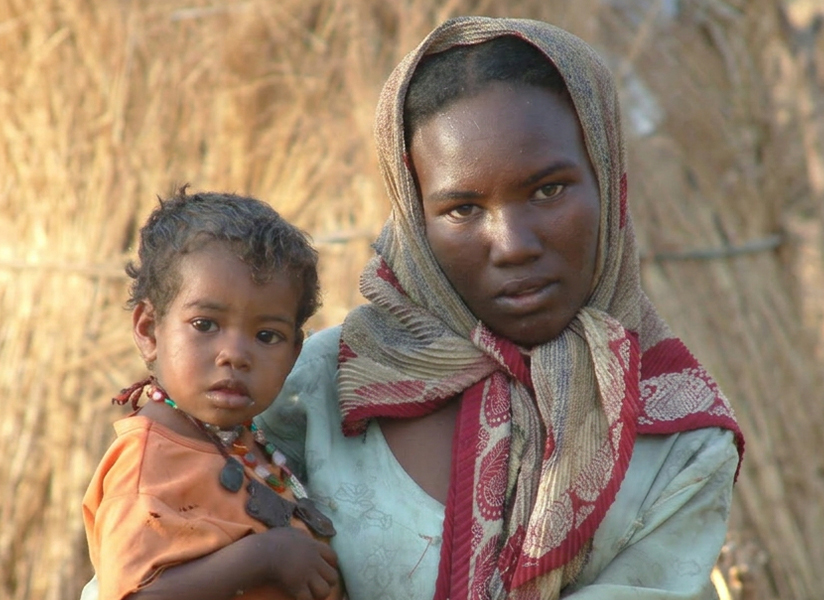 A young woman in a patterned headscarf holds a toddler in front of a straw-walled home.
