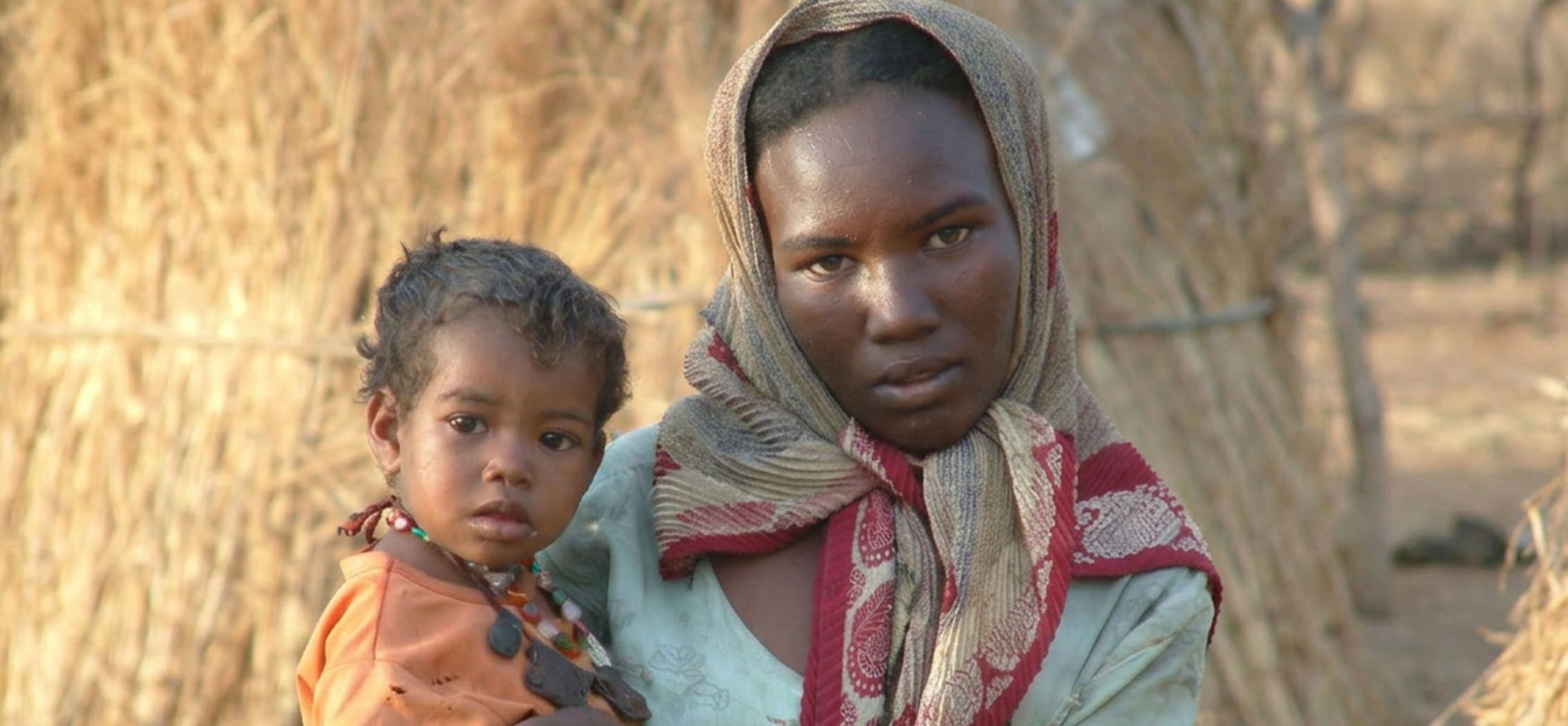 A young woman in a patterned headscarf holds a toddler in front of a straw-walled home.