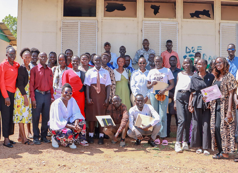 A large group of students, mentors, and parents pose together outside a school building, several holding certificates.