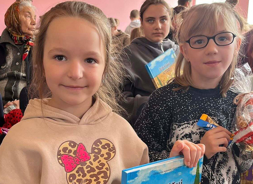 Two young girls smile at the camera in a crowded room where children hold gift bags.