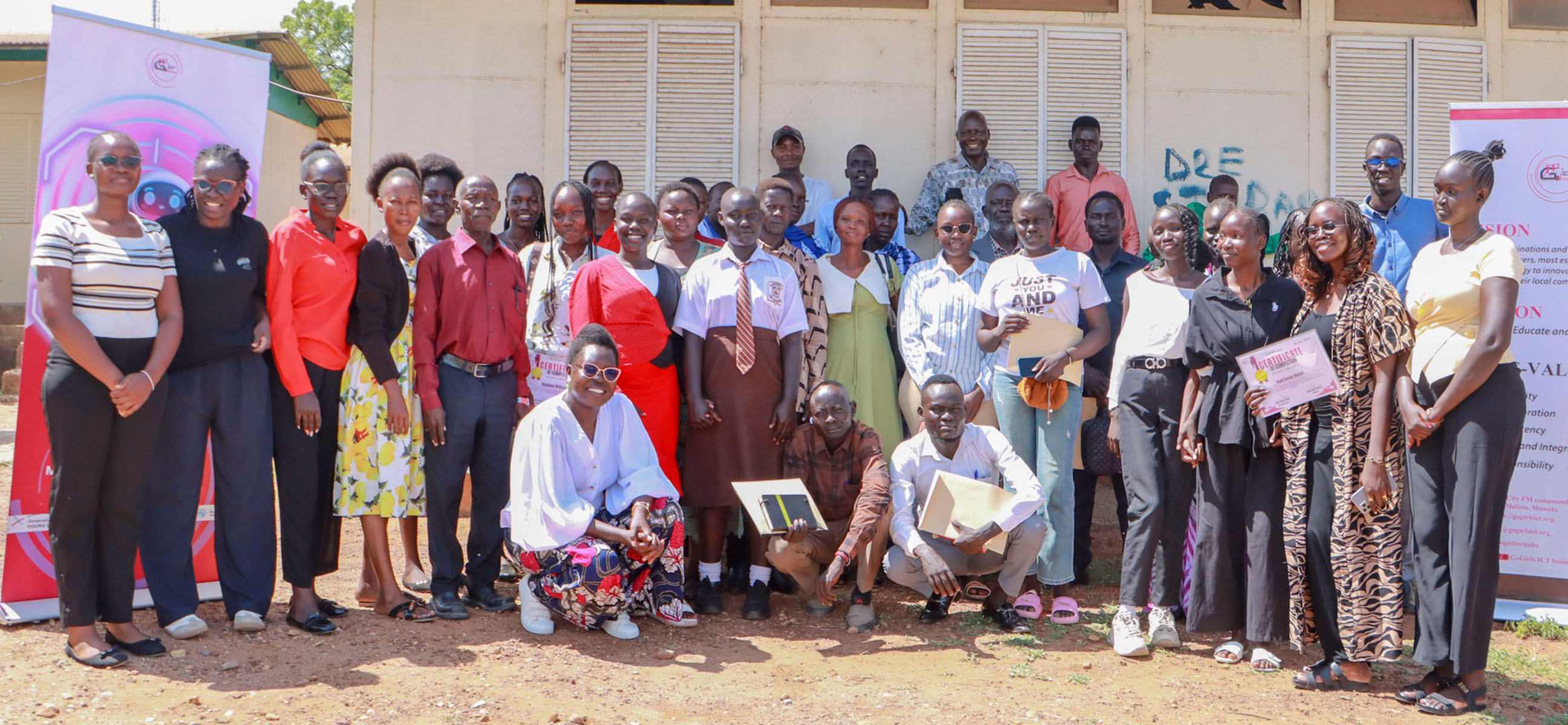 A large group of students, mentors, and parents pose together outside a school building, several holding certificates.