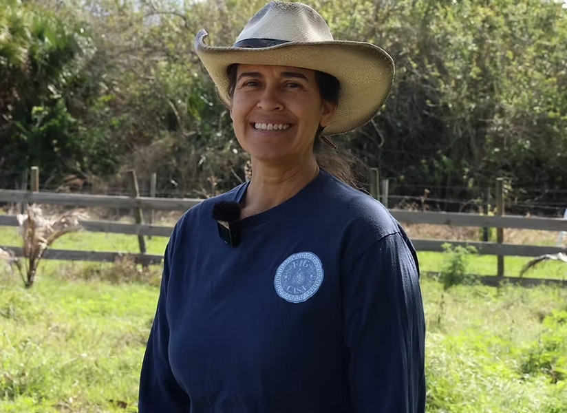 A smiling woman in a wide-brimmed hat stands in a green pasture beside a wooden fence.