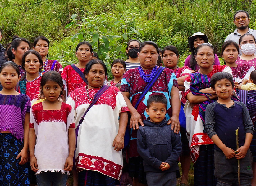 A large group of women and children, many in embroidered traditional blouses, stand together on a green hillside.