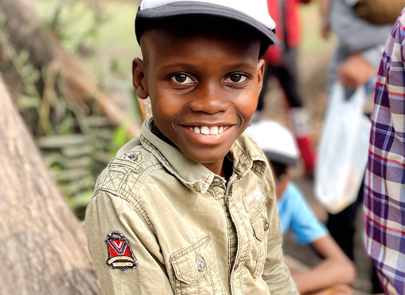 A smiling boy in a cap and khaki shirt looks at the camera at an outdoor gathering.