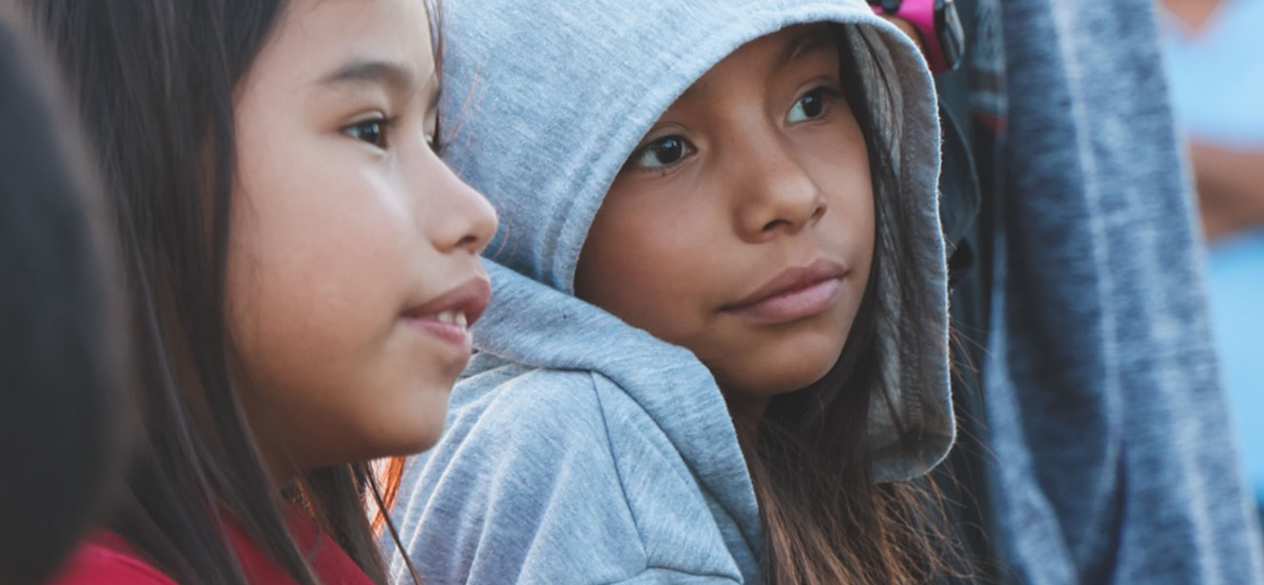 Two young girls, one in a gray hoodie, sit close together at an outdoor event.