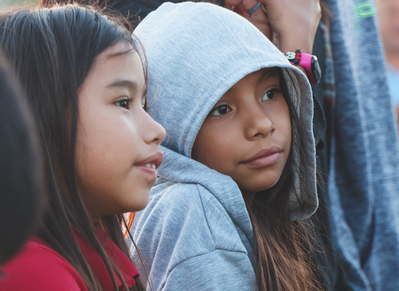 Two young girls, one in a gray hoodie, sit close together at an outdoor event.