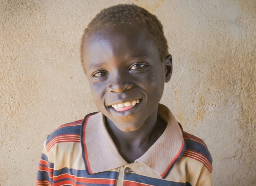 A smiling boy in a striped collared shirt stands against an earthen wall.