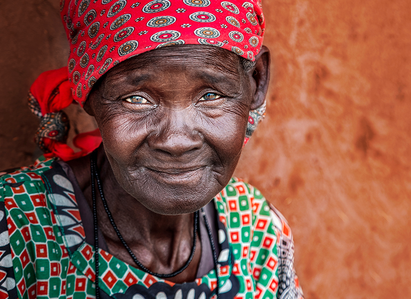 An elderly woman in a red headwrap smiles softly against a warm clay wall.