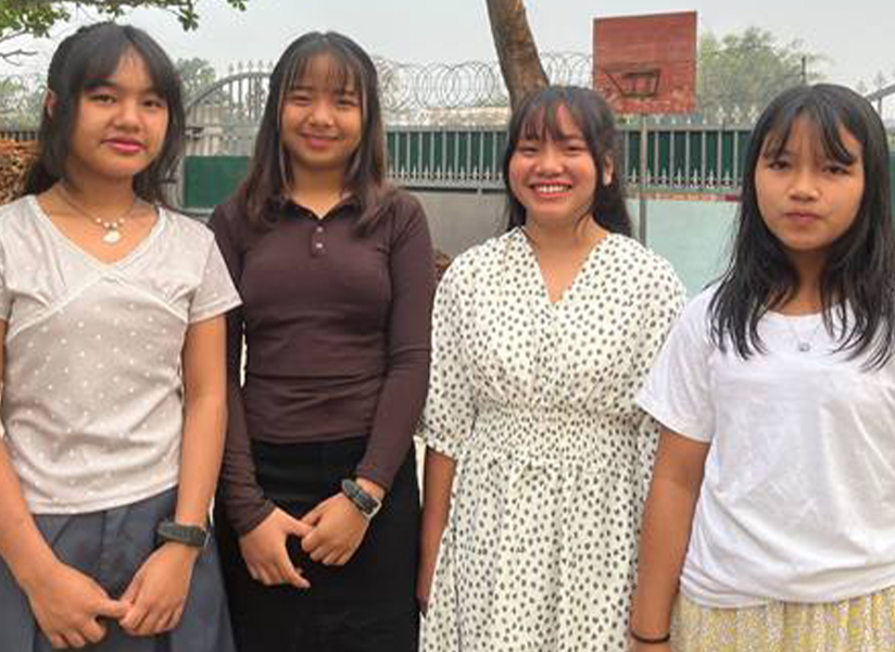 Four smiling teenage girls stand side by side in a courtyard.