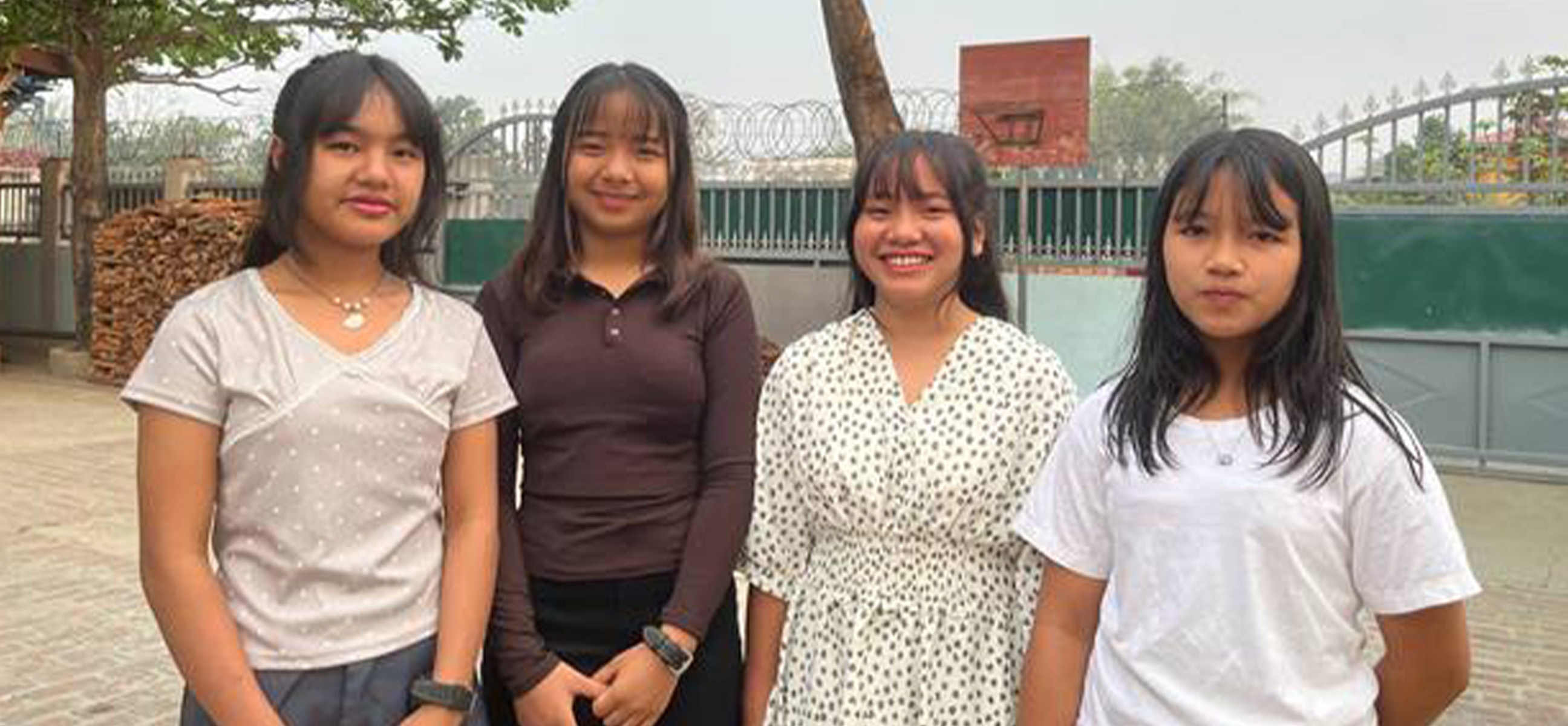 Four smiling teenage girls stand side by side in a courtyard.