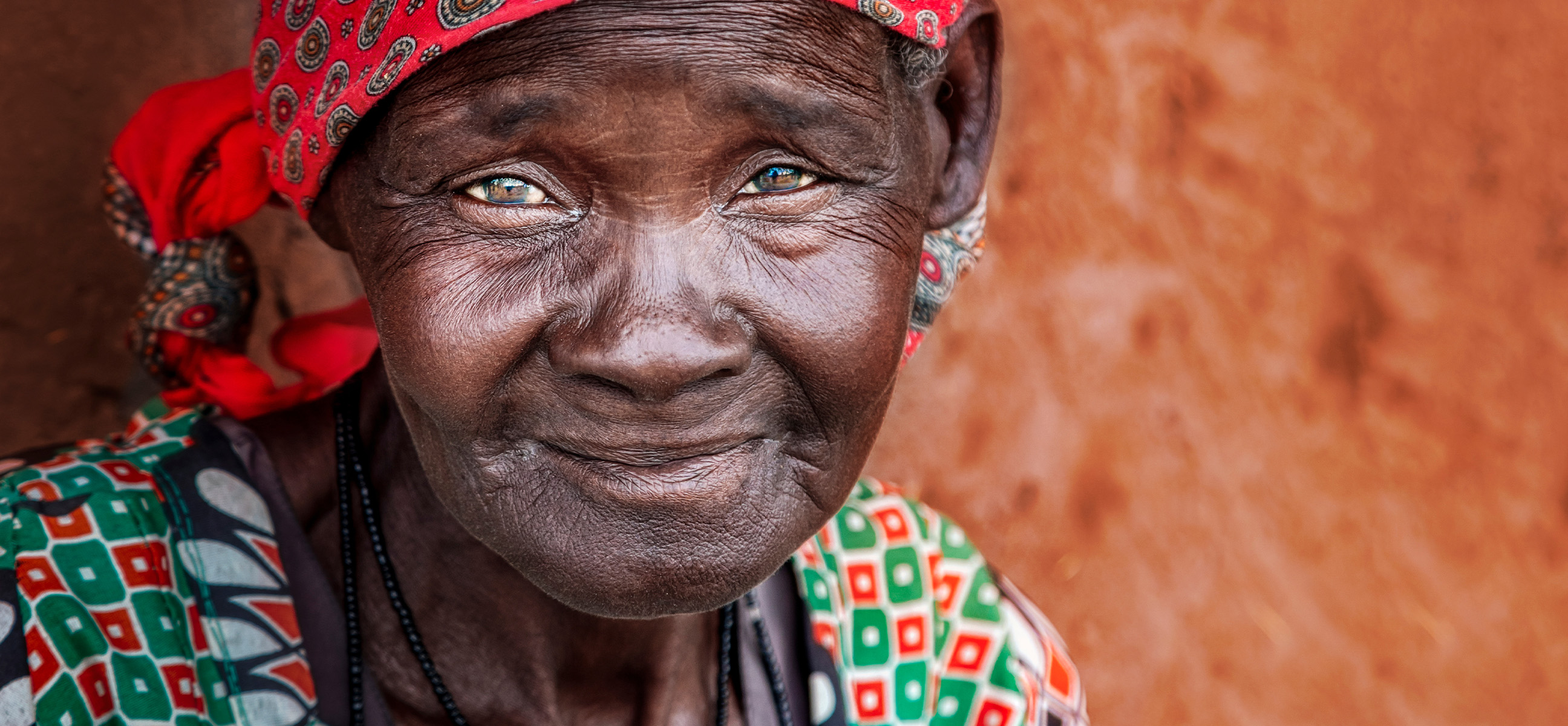 An elderly woman in a red headwrap smiles softly against a warm clay wall.