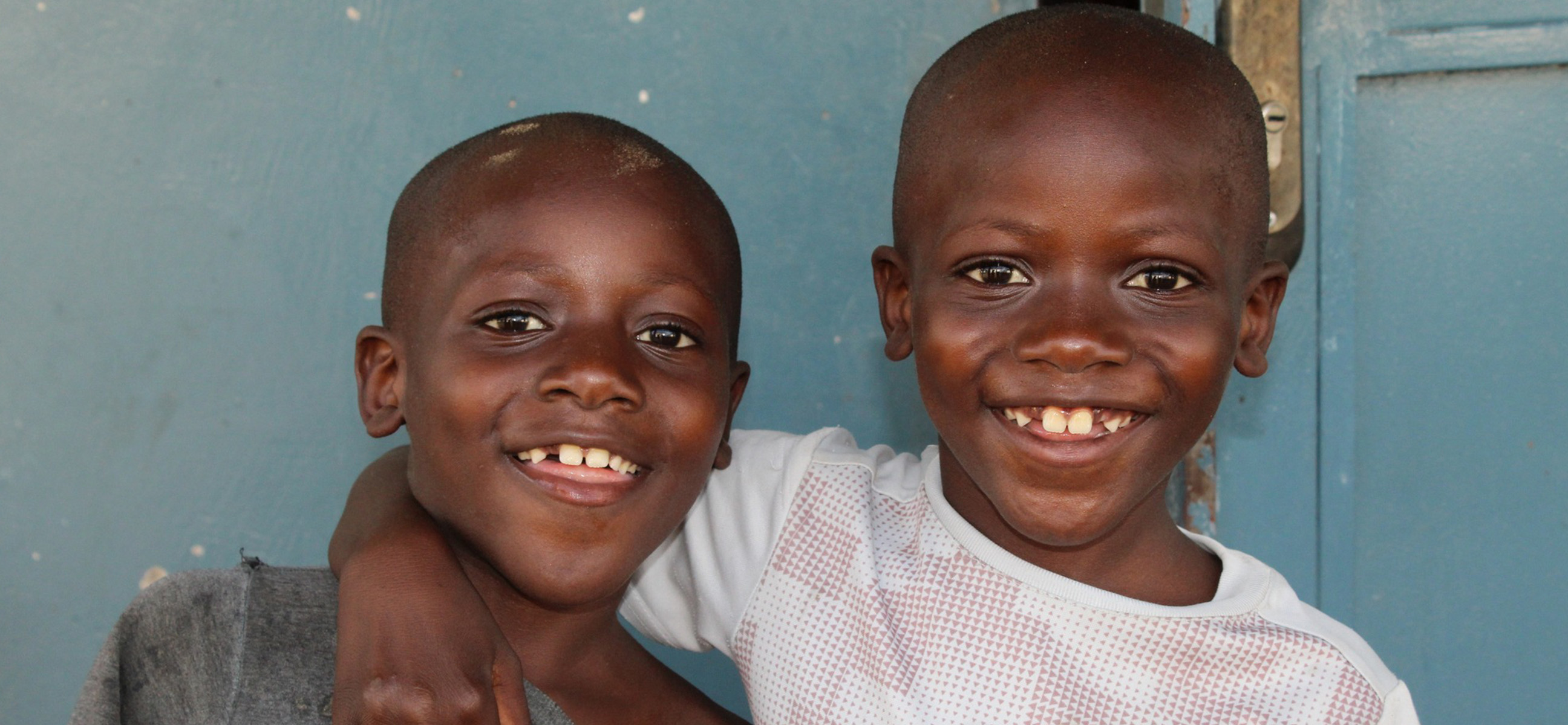 Two grinning young boys pose cheek to cheek in front of a blue wall.