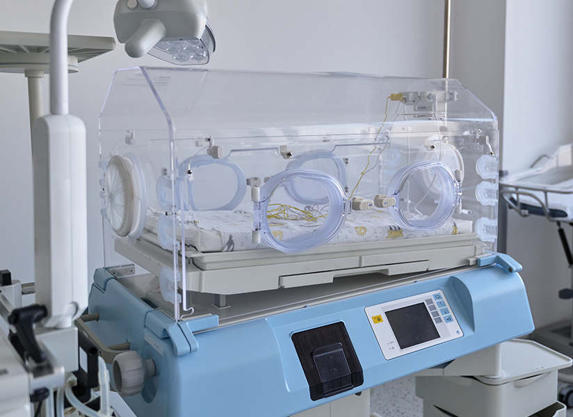A new infant incubator stands in a hospital neonatal ward beside empty bassinets.