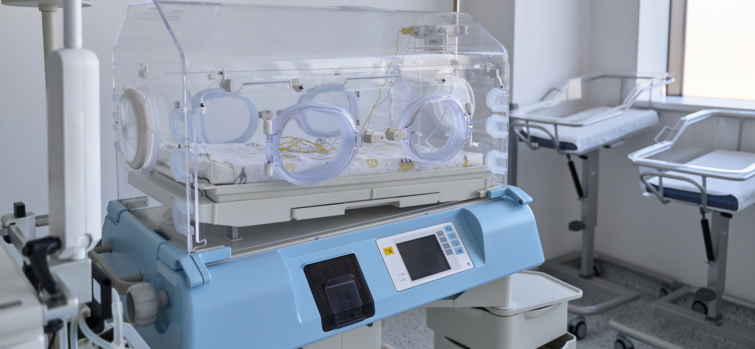 A new infant incubator stands in a hospital neonatal ward beside empty bassinets.