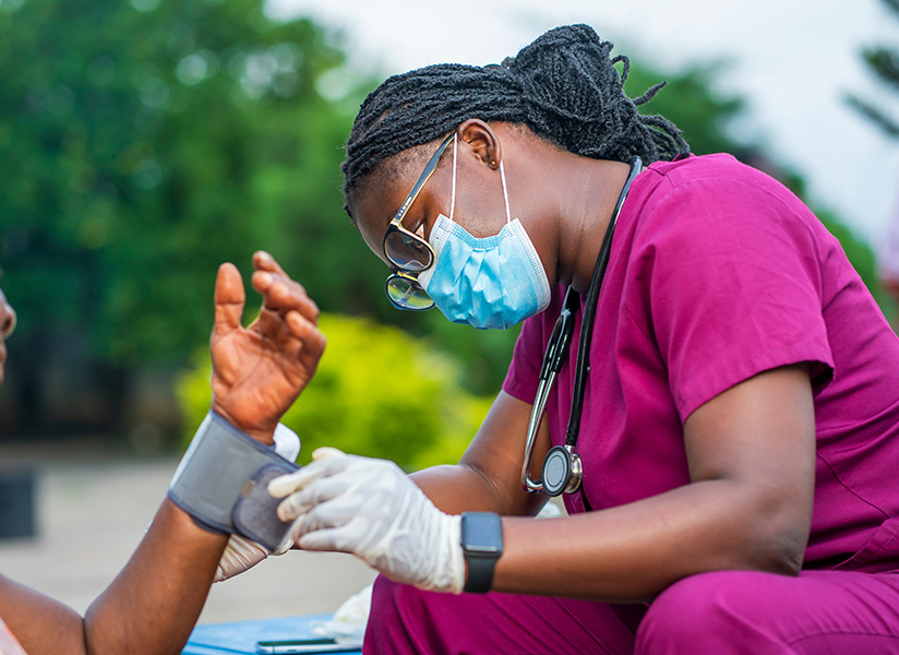 A health worker in magenta scrubs and a face mask checks an older patient's blood pressure outdoors.