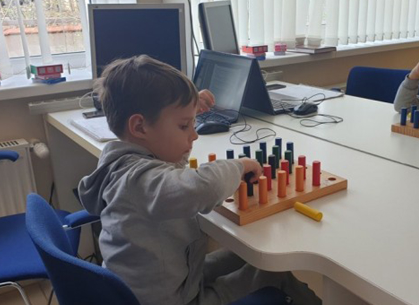 A young boy in a gray hoodie works on a colorful peg-board activity at a classroom table.