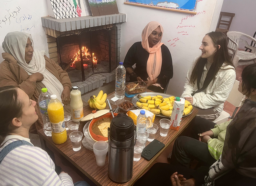 Women gather around a low table of food and drinks beside a glowing fireplace.