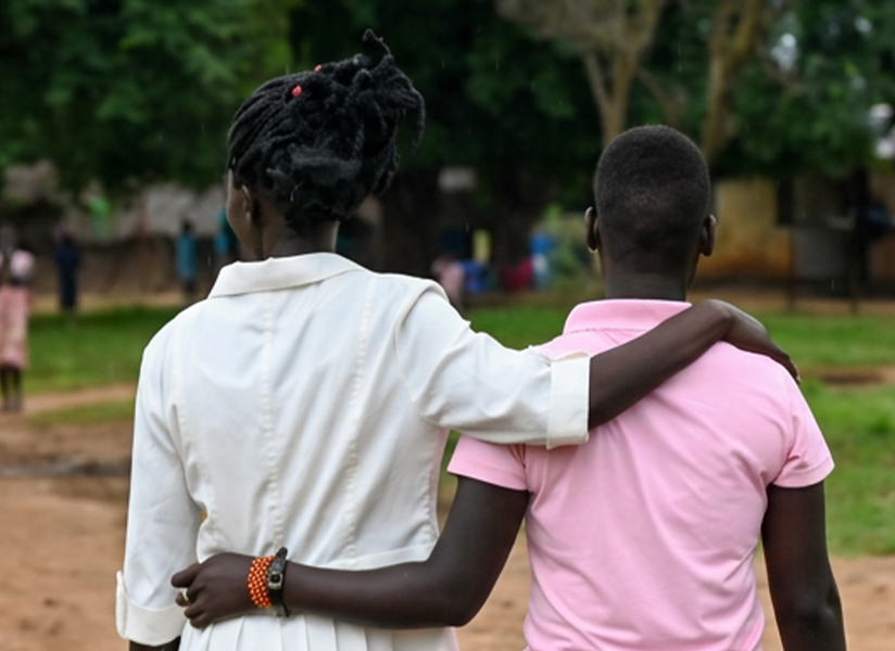 Seen from behind, two young people walk arm in arm down a dirt path toward a village.