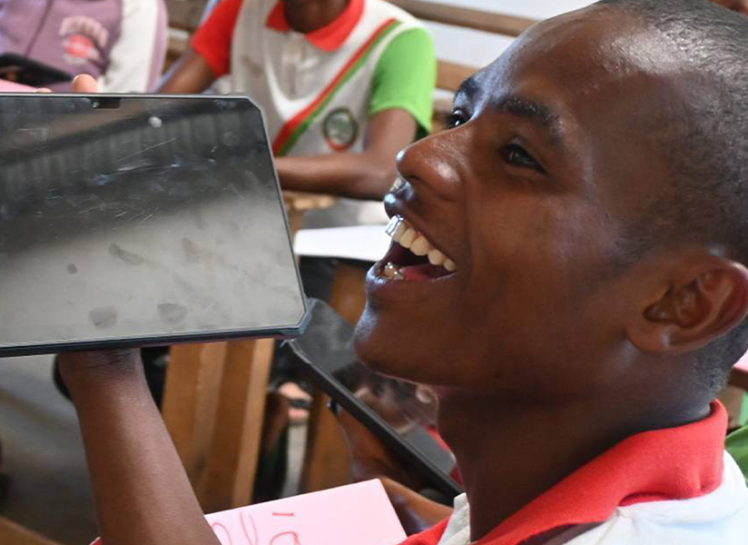 A laughing student holds up a tablet in a busy classroom.