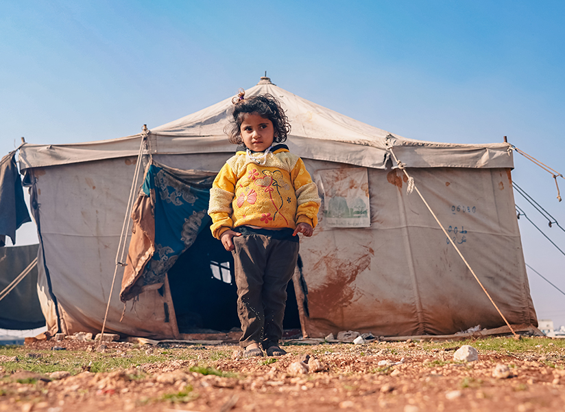 A young child in a yellow jacket stands in front of a weathered tent.