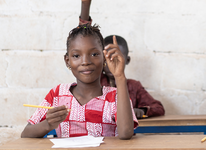 Smiling schoolgirls in colorful dresses raise their hands at their desks in a classroom.